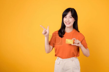 Foto de Alegre joven mujer asiática de 30 años con camisa naranja que sostiene la tarjeta de crédito y señala el dedo hacia el espacio libre de copia aislado en el fondo amarillo. positividad, compras y conceptos financieros. - Imagen libre de derechos