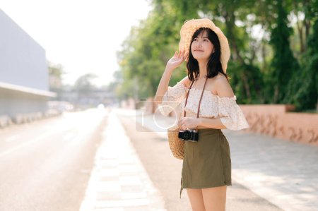 Foto de Portrait of young asian woman traveler with weaving hat and basket and a camera standing by the street. Journey trip lifestyle, world travel explorer or Asia summer tourism concept. - Imagen libre de derechos