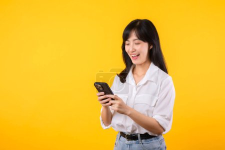 Téléchargez les photos : Portrait belle asiatique jeune femme sourire heureux portant chemise blanche et plantes en denim en utilisant smartphone isolé sur fond jaune studio. lire les nouvelles du concept de smartphone. - en image libre de droit