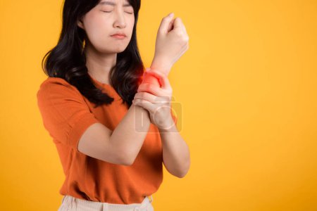 Foto de Destacando el cuidado de la salud de la artritis, mujer de 30 años asiática con camisa naranja mientras sostiene su muñeca con dolor. Ilustrando los desafíos de la artritis de muñeca, sirve como concepto para la salud y el bienestar. - Imagen libre de derechos