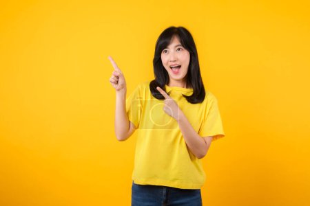 Foto de Capta la atención con este impresionante retrato de una joven asiática confiada que lleva una camiseta amarilla y vaqueros. Señala con el dedo el espacio libre para copiar. Perfecto para proyectos creativos. - Imagen libre de derechos