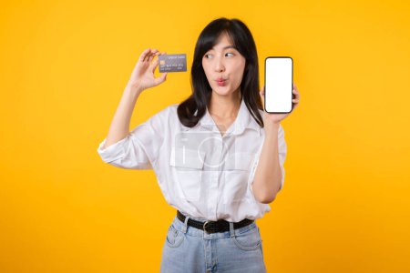 Téléchargez les photos : Portrait jeune femme asiatique portant une chemise blanche et un jean en jean avec un sourire heureux tenant une carte de crédit pour payer vos achats en ligne sur smartphone isolé sur fond jaune. shopping concept en ligne. - en image libre de droit