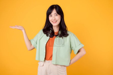 Foto de Vibración y alegría retrato joven mujer asiática lleva camisa verde pastel en camisa naranja y mostrar sonrisa feliz mientras señala gesto de la mano a espacio libre copia. Perfecto para añadir un toque animado a los proyectos. - Imagen libre de derechos