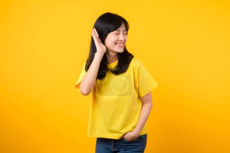 Photo for Capture attention portrait of Asian young woman. Wearing a yellow t-shirt and denim jeans, leans in to overhear and listen, evoking curiosity and intrigue. attention-grabbing promotions and discounts. - Royalty Free Image