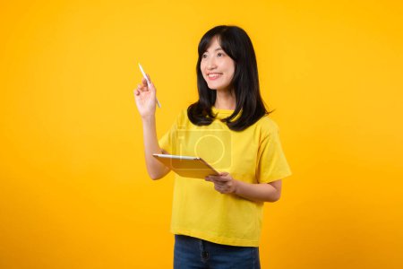 Téléchargez les photos : Jeune femme asiatique portant un t-shirt jaune et un jean montrant un sourire heureux tout en utilisant une tablette numérique, affichant une expression réfléchie et une idée créative. technologie de l'éducation concept de pensée innovant. - en image libre de droit