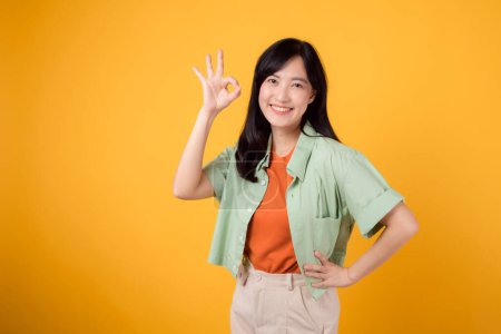 Téléchargez les photos : Confiance enveloppante et femme asiatique joyeuse de la trentaine portant une chemise verte et orange. Avec la main sur la hanche et le signe correct sur fond jaune, rayonne la positivité et l'assurance. Concept approuvé. - en image libre de droit