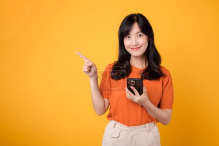 Téléchargez les photos : Ressentez la joie avec une jeune femme asiatique des années 30, portant une chemise orange, pointant vers l'espace libre de copie tout en utilisant un smartphone sur fond jaune. nouveau concept d'application. - en image libre de droit