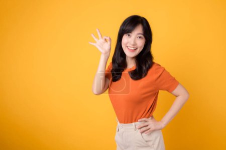 Foto de Encarnando positividad, la joven mujer asiática de 30 años hace alarde de estar bien con un signo de camisa naranja sobre fondo amarillo. Concepto de gesto de mano. - Imagen libre de derechos