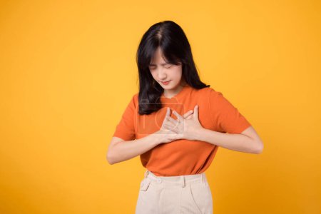 Foto de Mujer asiática joven atenta de 30 años, con una camisa naranja, sostiene las manos en el pecho sobre un fondo amarillo. enfermedad por ataque cardíaco, dolor torácico concepto de atención médica. - Imagen libre de derechos