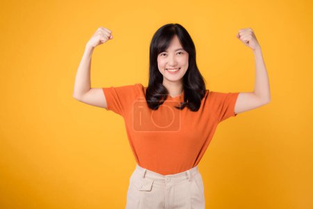 Téléchargez les photos : Confiante jeune femme asiatique trentaine célèbre avec un geste de signe de la main poing vers le haut, portant une chemise orange sur fond jaune. Concept d'autonomisation et de féminisme. - en image libre de droit