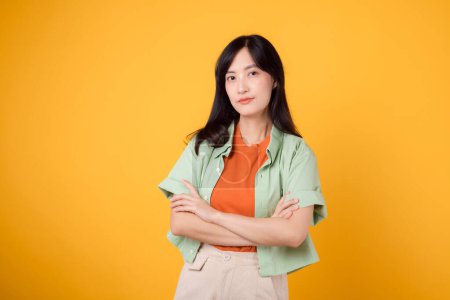 Foto de Abrace la confianza y el bienestar de la joven mujer asiática de 30 años con camisa naranja. muestra un gesto cruzado del brazo en el pecho contra el fondo amarillo, capturando un retrato de seguridad y autocuidado - Imagen libre de derechos