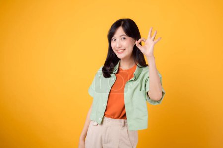 Téléchargez les photos : Charmante jeune femme asiatique des années 30, élégamment vêtue d'une chemise orange et d'un pull vert. Son geste captivant et son sourire doux brillent sur un fond jaune, concept de langage corporel. - en image libre de droit