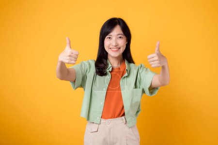 Photo for Young 30s Asian woman, elegantly attired in orange shirt and green jumper. Her thumbs up gesture, set against a yellow background, epitomizes the concept of affirmation. - Royalty Free Image