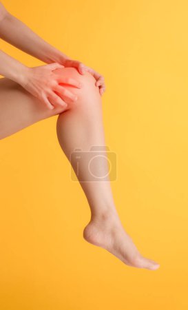 Téléchargez les photos : Abordez la douleur au genou et les soins de santé avec les mains de la femme tenant son genou douloureux sur fond jaune vif. Soulagement et concept de bien-être. - en image libre de droit
