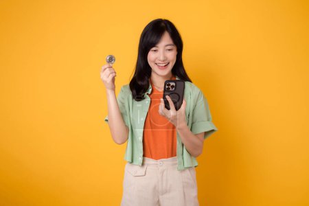 Foto de Innovación de las finanzas futuras con una joven mujer asiática vibrante de 30 años, vestida con camisa naranja y jersey verde, mostrando la pantalla del teléfono inteligente y la moneda criptográfica en el fondo amarillo - Imagen libre de derechos