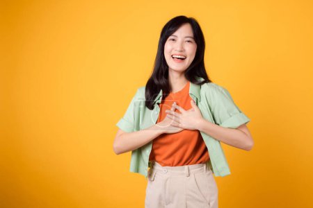 Téléchargez les photos : Soins de santé et bien-être avec une jeune femme asiatique trentaine, portant une chemise verte. Elle présente un geste de main sur sa poitrine sur un fond jaune, incarnant le concept de bien-être du corps. - en image libre de droit