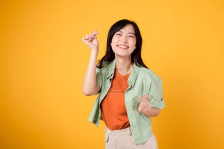Foto de Siente el amor en el aire con la encantadora joven mujer asiática de 30 años. Ella gentilmente muestra un mini gesto de corazón y una sonrisa suave. concepto de lenguaje corporal. con una camisa naranja y un jersey verde. - Imagen libre de derechos