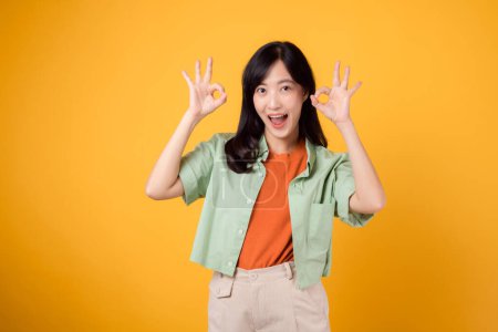 Foto de Joven mujer asiática de 30 años, elegantemente vestida con camisa naranja y jersey verde. Su gesto de mano bien y su sonrisa suave, capturados sobre fondo amarillo, transmiten un mensaje conmovedor a través del lenguaje corporal. - Imagen libre de derechos