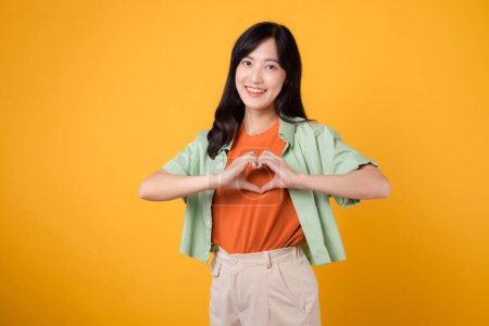 Foto de Salud y bienestar como mujer asiática de 30 años, con una camisa verde, muestra un gesto de la mano del corazón en su pecho contra un fondo amarillo vibrante. Experimente la encarnación del bienestar corporal - Imagen libre de derechos