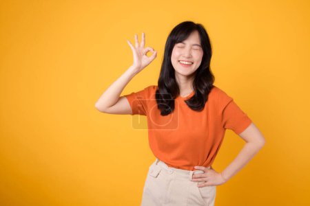 Photo for Expressing positivity, 30s Asian woman wears orange shirt, exhibits okay sign on vibrant yellow background. Hands gesture concept. - Royalty Free Image