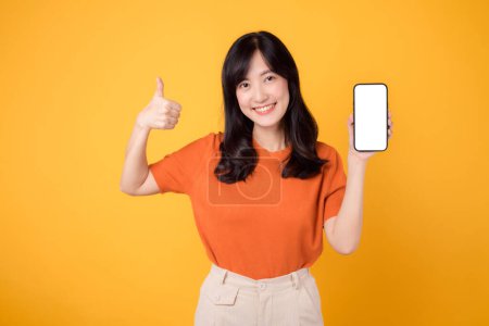 Foto de Mujer asiática joven de 30 años con camisa naranja que muestra el pulgar hasta la pantalla de visualización en blanco del teléfono inteligente aislado en el fondo amarillo. alegre cara feliz persona femenina con nuevo concepto de tecnología de aplicación. - Imagen libre de derechos