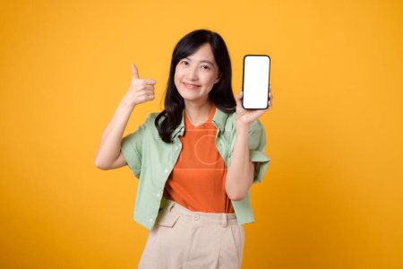 Téléchargez les photos : Nouvelle application mobile avec une jeune femme asiatique joyeuse trentaine, portant une chemise orange et un pull vert, affichant un écran de smartphone avec un geste pouce vers le haut sur fond de studio jaune. - en image libre de droit