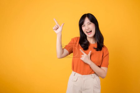 Foto de Encarna alegría con una joven asiática de 30 años, vistiendo una camisa naranja, señalando espacio libre para copiar sobre fondo amarillo. recomendar el concepto de producto. - Imagen libre de derechos