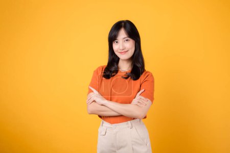 Téléchargez les photos : Embrassez les vibrations confiantes avec une jeune femme asiatique trentaine, portant une chemise orange, présentant un geste de signe du bras croisé sur fond jaune. - en image libre de droit