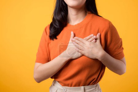 Téléchargez les photos : Gros plan de la jeune femme asiatique trentaine, portant une chemise orange, tient la main sur la poitrine sur fond jaune. concept de bien-être. - en image libre de droit