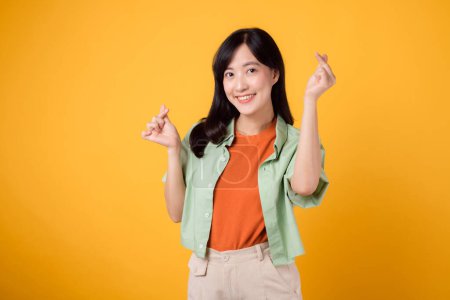 Photo for Warmth of affection and happiness with young 30s Asian woman, dressed in an orange shirt and green jumper. Her mini heart gesture and gentle smile express a profound message through body language. - Royalty Free Image