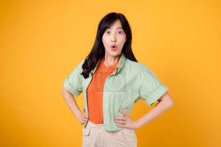 Foto de Expresiva y sorprendente, una joven mujer asiática de 30 años con una camisa verde en una camisa naranja que muestra la cara sorpresa mientras está de pie con los brazos akimbo. inesperada con imagen vibrante sobre fondo amarillo. - Imagen libre de derechos