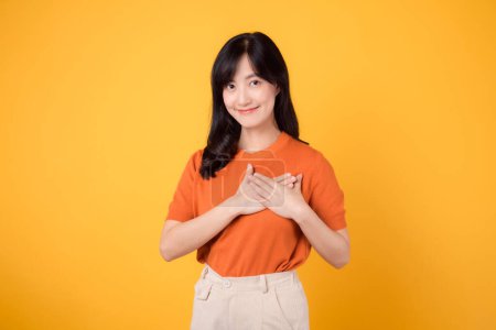 Téléchargez les photos : Joyeuse jeune femme asiatique trentenaire, portant une chemise orange, tient la main sur la poitrine sur fond jaune. concept de bien-être. - en image libre de droit