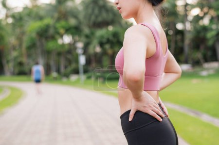 Foto de Una corredora. Mujer asiática joven de 30 años que usa ropa deportiva rosa sosteniendo su dolor de espalda y cadera después de hacer ejercicio en el parque público. Concepto de dolor en la actividad. - Imagen libre de derechos