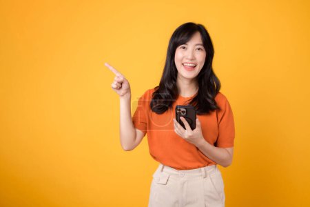 Téléchargez les photos : Répandre le bonheur avec une jeune femme asiatique trentaine, revêtant une chemise orange, pointant vers l'espace libre de copie tout en utilisant un smartphone sur fond jaune. nouveau concept d'application. - en image libre de droit