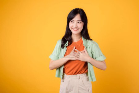 Téléchargez les photos : Embrassez l'amour des affaires avec une jeune femme asiatique trentaine portant une chemise verte sur une chemise orange, rayonnant de bonheur. Concept de promotion de l'accord d'amour dans un contexte jaune vibrant. - en image libre de droit
