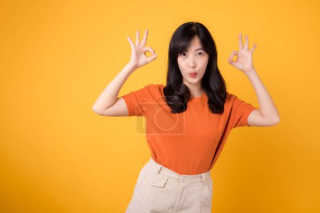 Foto de En un vibrante telón de fondo amarillo, una joven mujer asiática de 30 años, vestida con una camisa naranja, muestra el gesto de señal aceptable. Concepto de gesto de manos. - Imagen libre de derechos