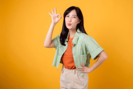 Foto de Joven mujer asiática de 30 años, vistiendo una camisa naranja y jersey verde. Su gesto de mano bien y su sonrisa suave, aislada sobre un fondo amarillo vibrante, exudan positividad a través del lenguaje corporal. - Imagen libre de derechos