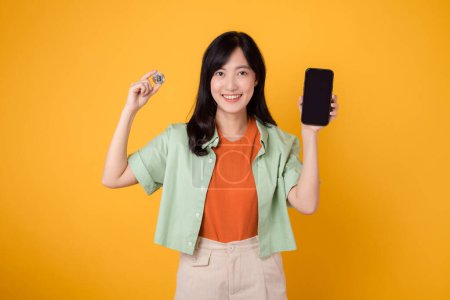Foto de El futuro de las finanzas con una cautivadora joven mujer asiática de 30 años, elegantemente vestida con camisa naranja y jersey verde, utilizando la pantalla del teléfono inteligente y la moneda criptográfica sobre fondo amarillo. - Imagen libre de derechos