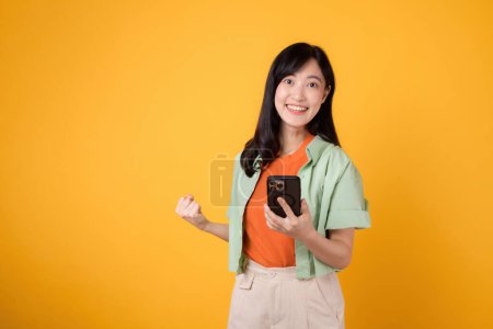 Téléchargez les photos : Jeune femme asiatique excitée des années 30, vêtue d'une chemise orange et d'un pull vert, utilisant un smartphone avec un geste poing vers le haut sur fond jaune. Concept mobile avec de bonnes nouvelles. - en image libre de droit