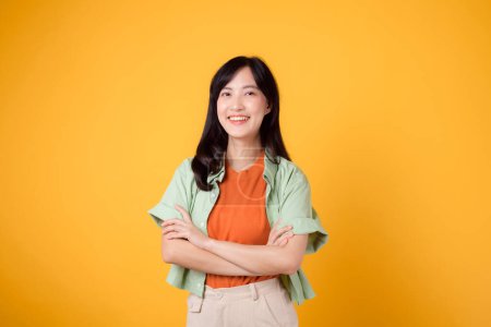 Foto de Confianza y bienestar con una joven mujer asiática de 30 años con una camisa naranja. Su gesto cruzado del brazo en su pecho contra un fondo amarillo vibrante emana seguridad en sí misma y paz interior. - Imagen libre de derechos