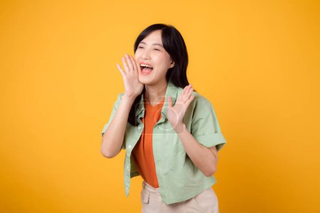Photo for Energetic young Asian woman 30s wearing a green shirt on an orange background, shouting with excitement. Explore the concept of discount shopping promotion with this vibrant image. - Royalty Free Image