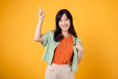 Téléchargez les photos : Débloquez des économies et magasinez maintenant ! jeune femme asiatique trentaine, portant une chemise verte sur une chemise orange. visage heureux et doigt pointeur pour libérer de l'espace de copie sur fond jaune vibrant. - en image libre de droit