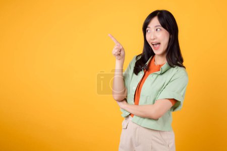 Téléchargez les photos : Jeune femme asiatique trentaine, portant une chemise verte sur une chemise orange. visage heureux et doigt pointeur pour libérer de l'espace de copie sur fond jaune vif. Débloquez des économies et magasinez maintenant! - en image libre de droit