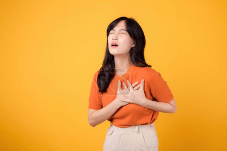 Foto de Mujer asiática preocupada de 30 años, vistiendo una camisa naranja, sostiene las manos en el pecho sobre un fondo amarillo. enfermedad por ataque cardíaco, dolor torácico concepto de atención médica. - Imagen libre de derechos