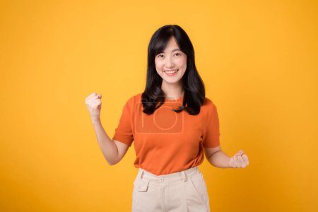 Téléchargez les photos : Embrassez la confiance comme une jeune femme asiatique trentaine lève le poing vers le haut signe geste, portant une chemise orange sur fond jaune. - en image libre de droit