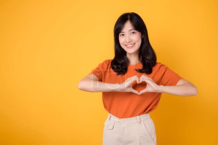 Photo for Healthcare and wellness embodied by an Asian woman 30s. Experience body wellness as she showcases a heart hand gesture on her chest against a vibrant yellow background - Royalty Free Image
