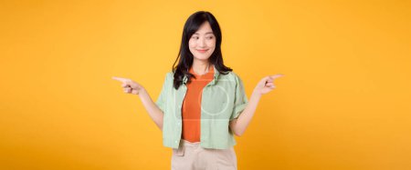Photo for Best deal promotion concept featuring young 30s Asian woman, wearing green shirt on orange shirt. With happy face, points her finger to free copy space, choose deals offers against yellow backdrop. - Royalty Free Image