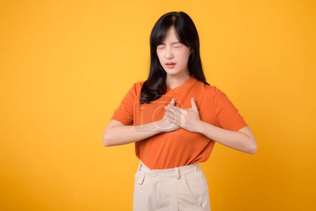 Photo for Caring young Asian woman 30s, wearing an orange shirt, holds hands on chest on yellow background. heart attack disease, chest pain health care concept. - Royalty Free Image