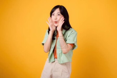 Energetic Asian woman 30s in a vibrant green and orange shirt, shouting with enthusiasm. Explore the concept of discount shopping promotion with this isolated yellow background photo.
