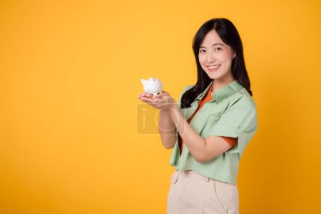 Photo for Happy young Asian woman 30s, wearing orange shirt and green jumper, showcases piggy bank on yellow background. Financial money concept. - Royalty Free Image